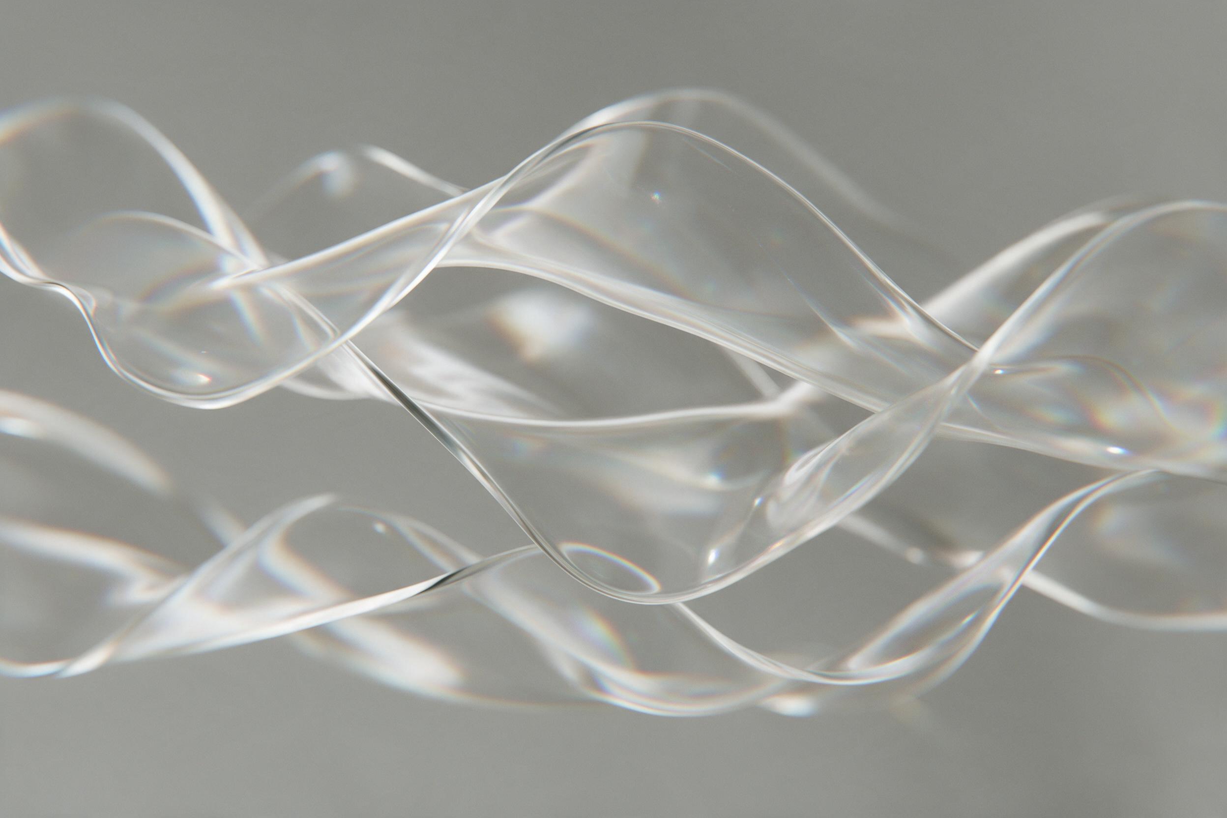 A minimalist composition of fluid, translucent glass waves suspended in mid-air, refracting soft morning light into subtle rainbow prisms across a muted gray backdrop. The flowing shapes overlap with delicate precision, their edges blurring gently with a shallow depth of field, creating a serene, me