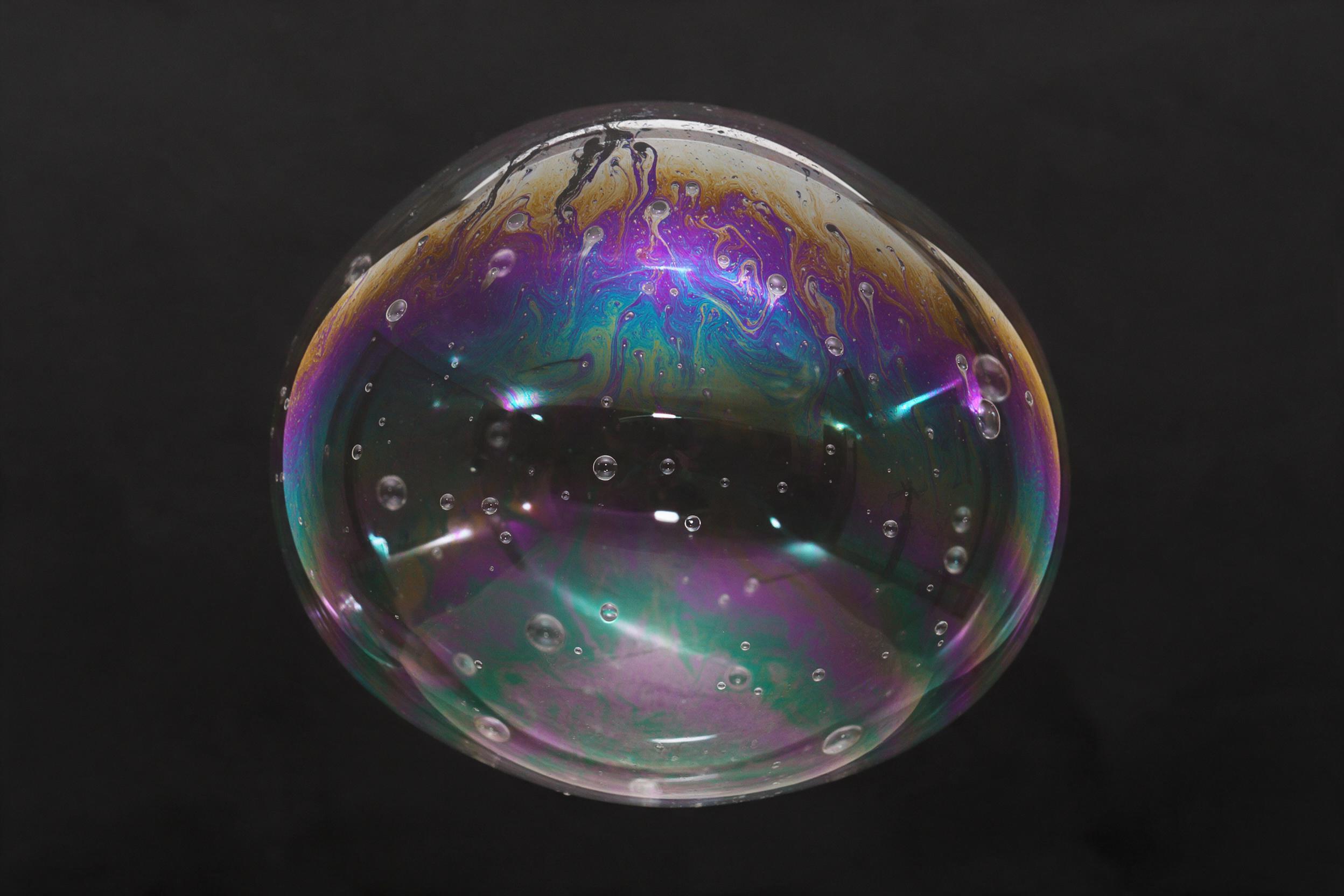 Macro shot of iridescent soap film, swirling liquid gradients in violet, teal, and amber, delicate bubbles and caustic light patterns, soft studio diffused lighting, minimal composition against matte black background