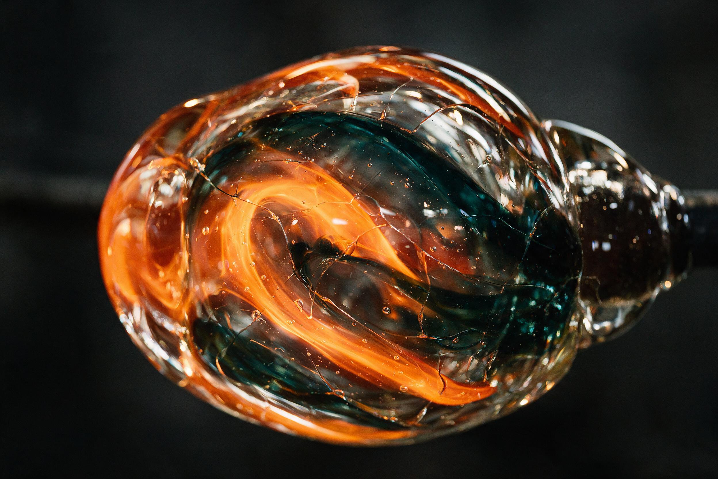 Narrow macro shot of molten glass being shaped, glowing orange and deep teal swirls trapped in transparent layers, fine bubbles and sharp cracks catch warm side light, dark charcoal background, shallow depth of field with slight blur at edges, smooth high-contrast textures, no text