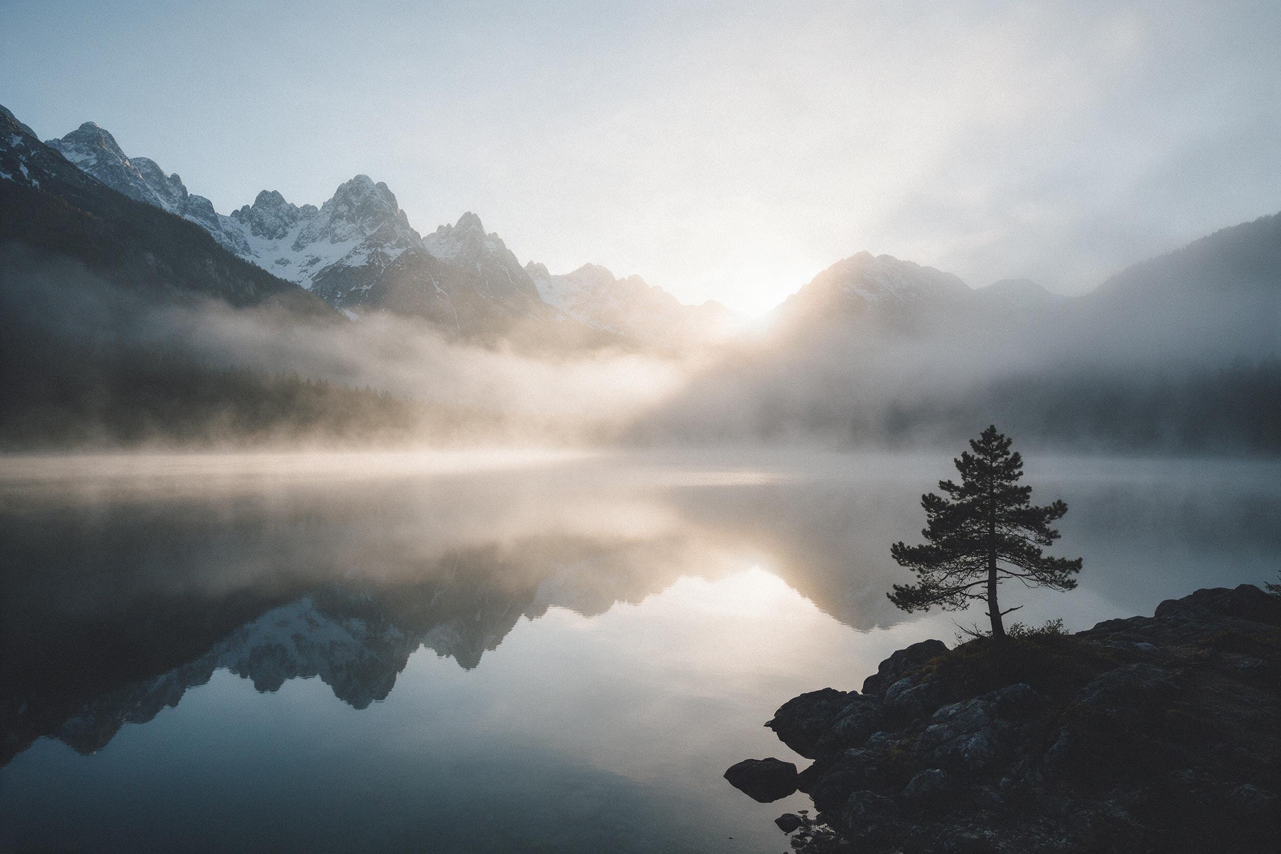 Misty alpine lake at dawn, glassy water reflecting jagged snow-capped peaks, soft golden light spilling over ridgelines through wispy fog, a lone pine silhouetted on rocky shore, fine grain film aesthetic, subtle atmospheric haze, wide-angle composition leading eye to horizon glow