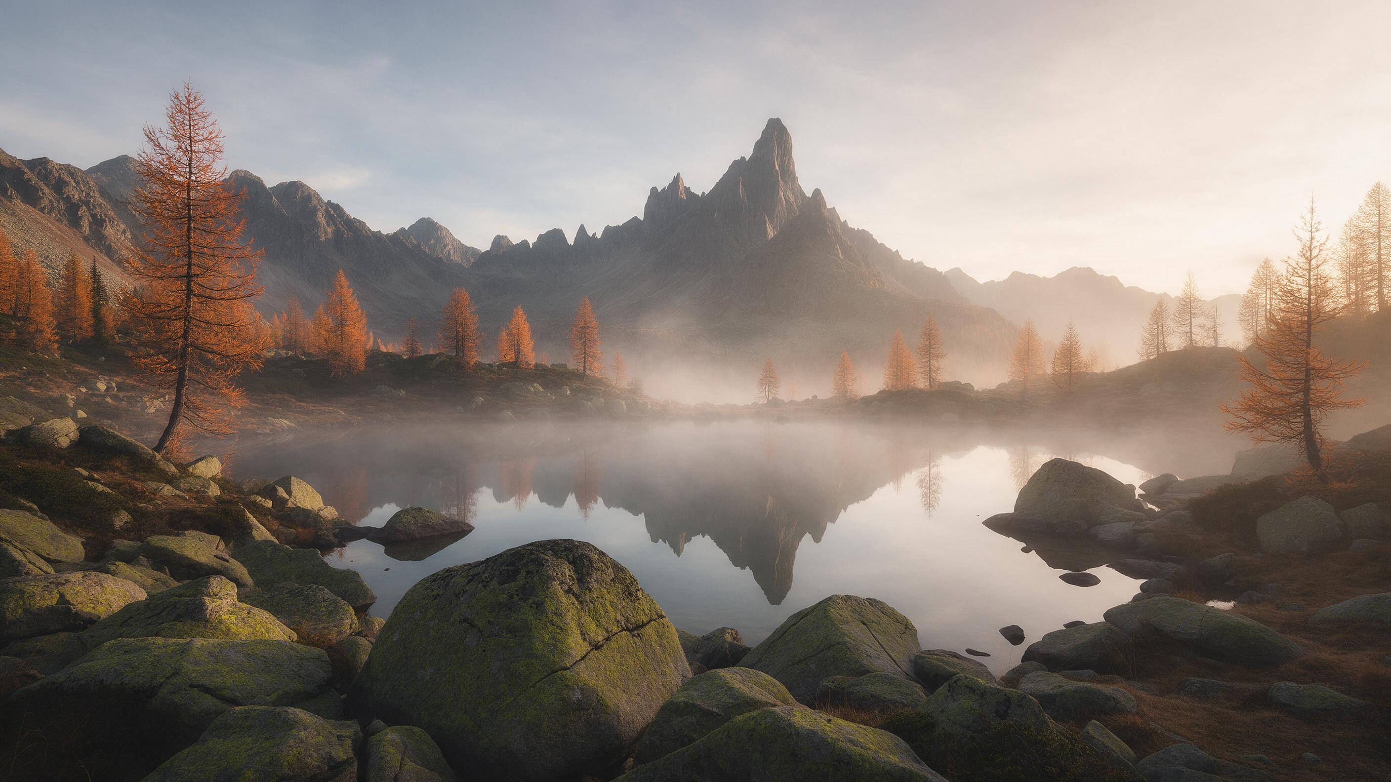 Golden hour over a misty alpine lake, jagged peaks reflected in still water, scattered autumn larches glowing orange, soft warm light, gentle haze, foreground boulders with moss, wide-angle composition, natural colors, no people.