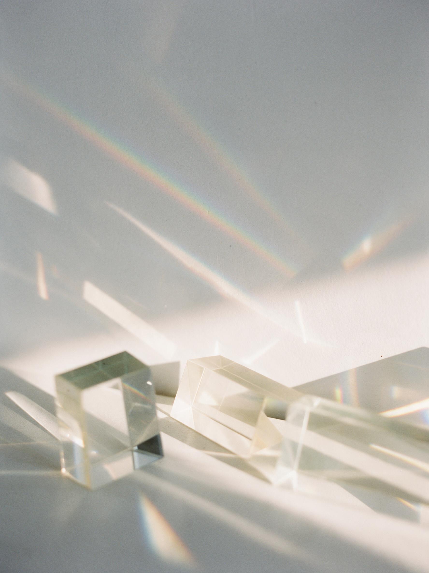 A minimalist composition of translucent glass prisms refracting warm afternoon sunlight into subtle rainbow bands across a smooth white plaster wall, with soft shadows and gentle geometric interplay, shot on medium format film with natural grain and muted tones