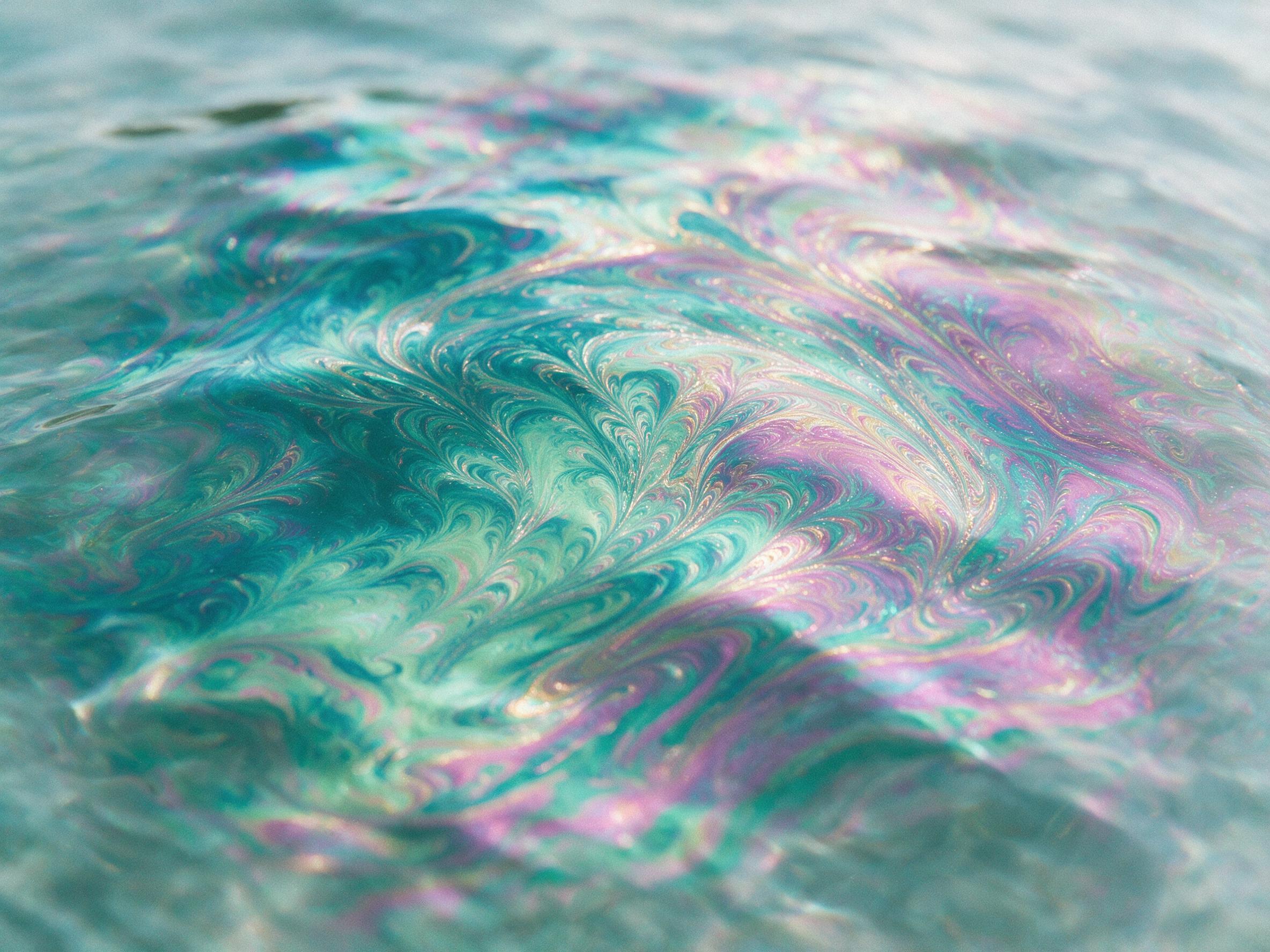 A macro shot of swirling iridescent oil film on water, delicate fractal patterns in teal and magenta, soft diffused daylight from above, gentle ripples distorting reflections, shallow depth of field, smooth gradients, subtle grain, no text