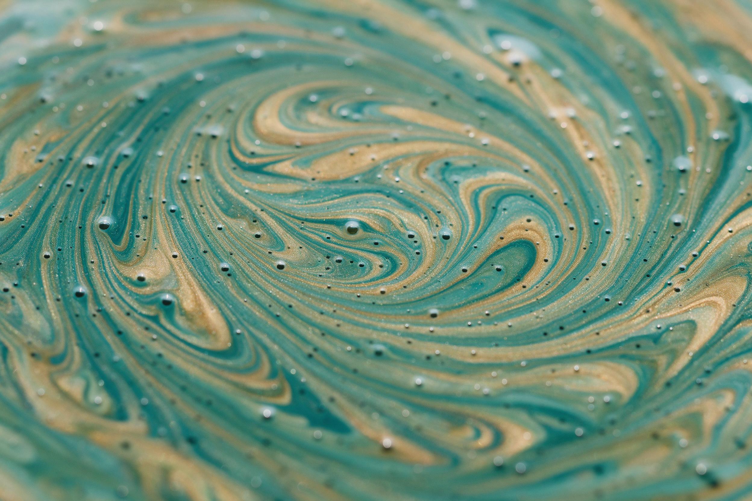Abstract macro shot of swirling liquid paint, teal and gold ribbons interweaving with microscopic air bubbles, wet glossy surface reflecting scattered skylight, 50mm lens close-up, shallow depth of field, fine texture detail, natural color palette