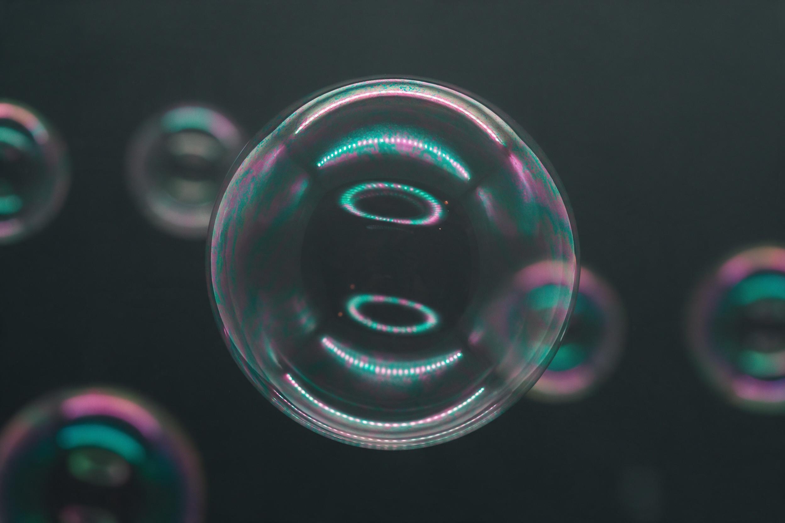 A macro shot of iridescent soap bubbles floating against a dark charcoal background, each sphere reflecting distorted concentric light rings in teal and magenta, fine liquid film texture with subtle color interference patterns, shallow depth of field, soft studio rim lighting, gentle haze at edges