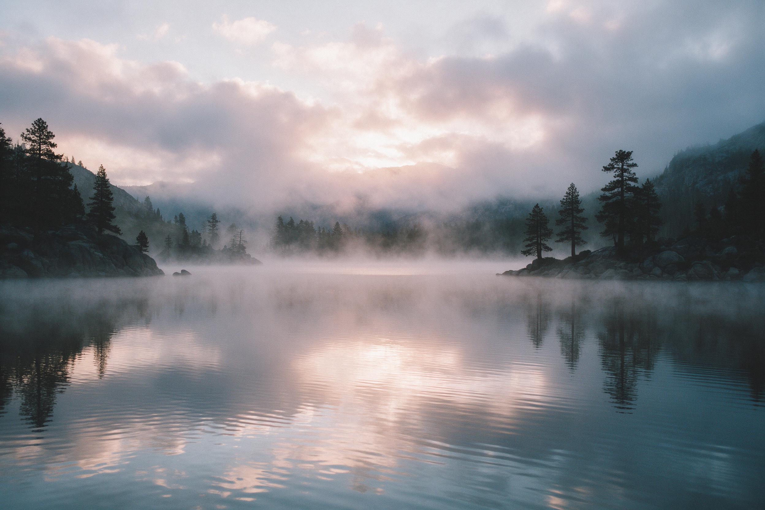 A serene mountain lake at dawn, mist curling over glassy water, silhouettes of pine trees on rocky shores, soft pink and gold light breaking through cloud layers, gentle ripples reflecting the sky, quiet atmosphere with no human presence, natural color grading, subtle film grain.