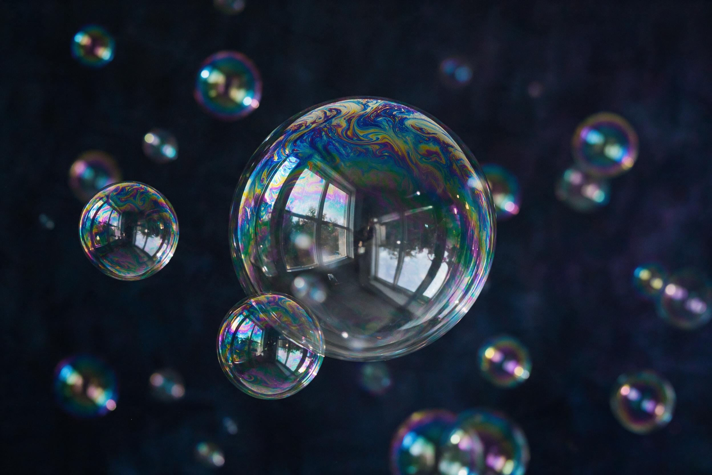 A close-up of iridescent soap bubbles floating against a dark matte background. Thin film distortion creates swirling rainbow patterns. Soft, diffused side lighting, shallow depth of field with few bubbles sharp, others blurred. Subtle reflection of a window frame visible in the largest bubble. Slig