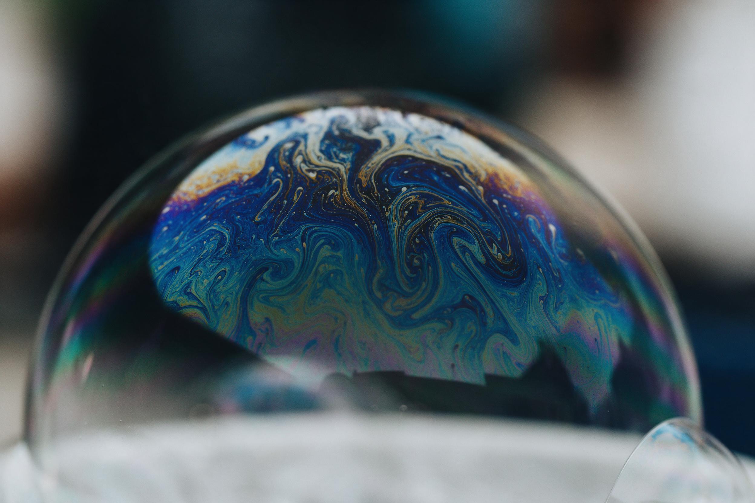 Macro shot of iridescent soap film, swirling liquid patterns with deep blues, teals, and gold, soft diffused light from side, subtle bokeh background, minimal composition, fine texture details