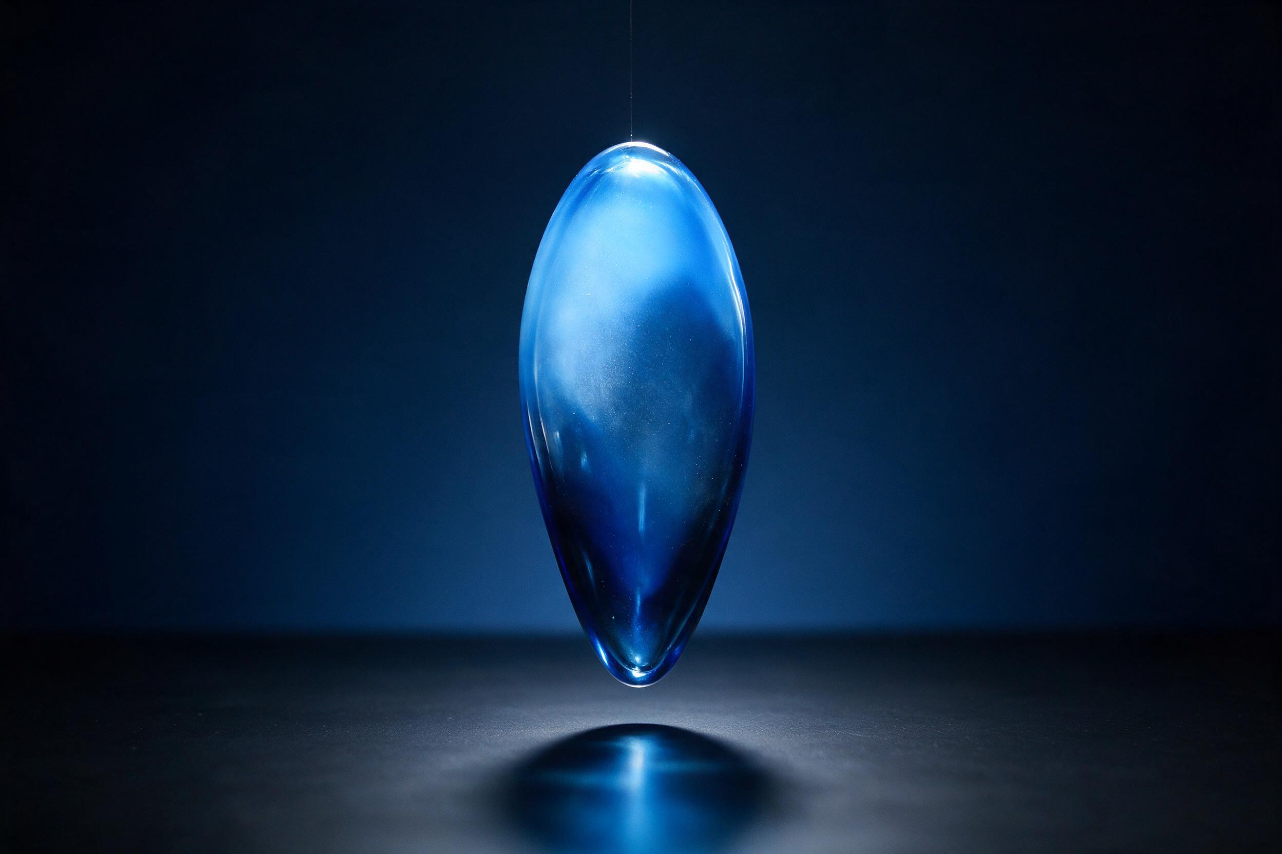 A single luminous teardrop-shaped glass sculpture suspended in mid-air, illuminated by a soft blue gradient light, with subtle reflections on a dark matte surface, minimal composition, smooth curves, slight grain, natural studio lighting.