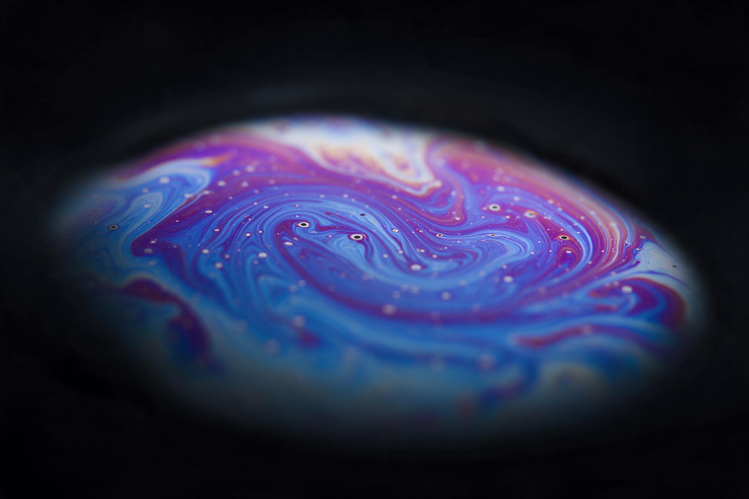 Abstract macro photography of swirling oil and water emulsion, iridescent blues and violets, delicate bubbles scattering light, soft studio reflection, blurred edges fading to deep black, silky smooth texture, warm magenta accents, minimalist composition