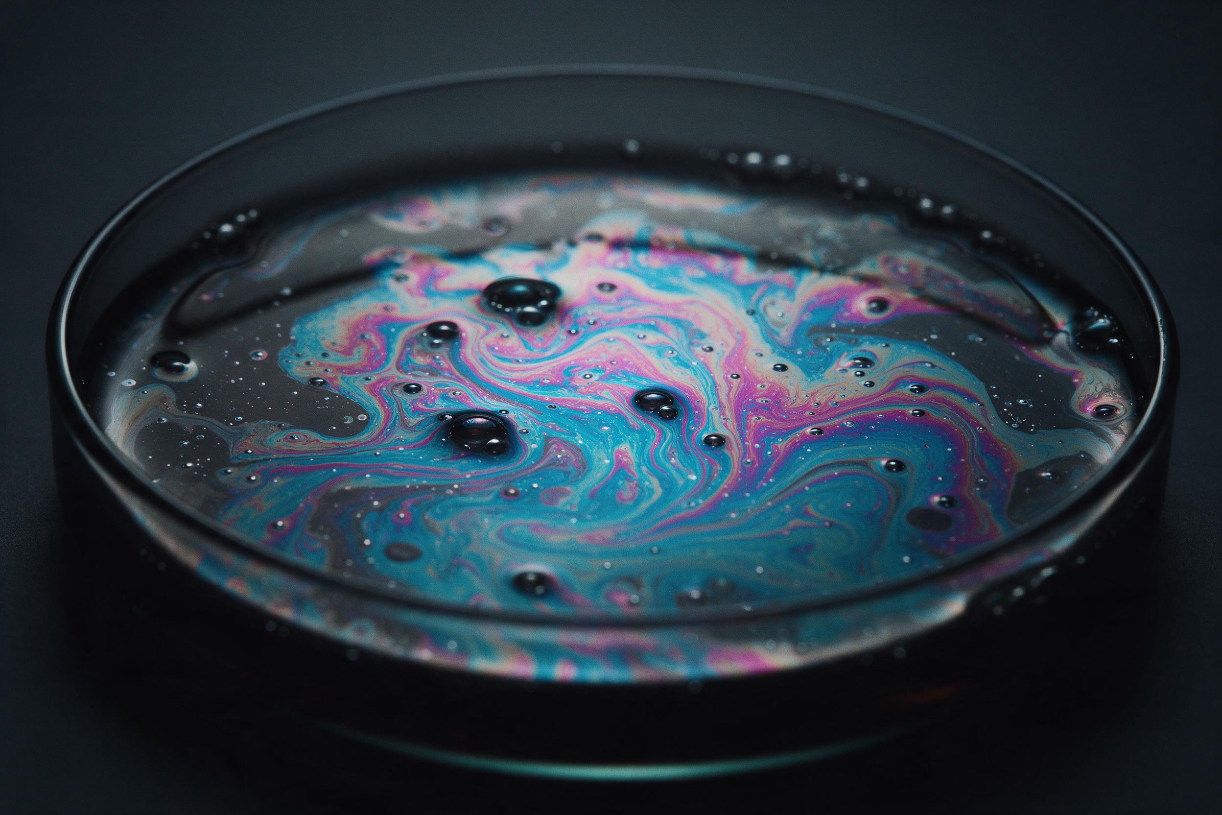 Macro shot of swirling oil and water in a dark petri dish, iridescent cyan and magenta blooms, delicate bubbles catching soft studio light, shallow depth of field, gentle reflections, fine grain.