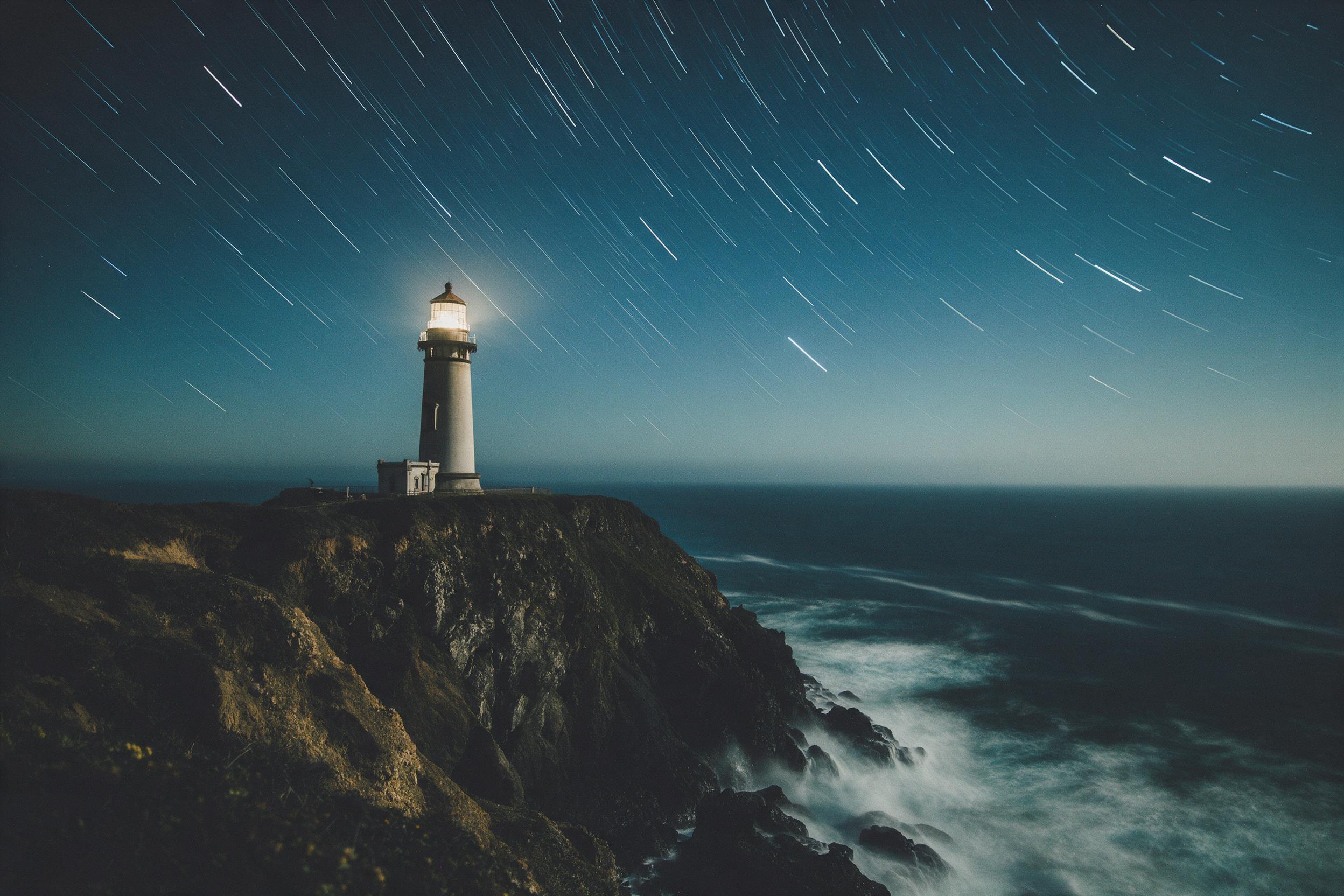 A solitary lighthouse on a rugged cliff, starlit sky, ocean waves crashing below, long-exposure light trails from stars, deep blue and teal tones, tranquil yet dramatic mood, fine grain film aesthetic, wide angle composition with lighthouse off-center