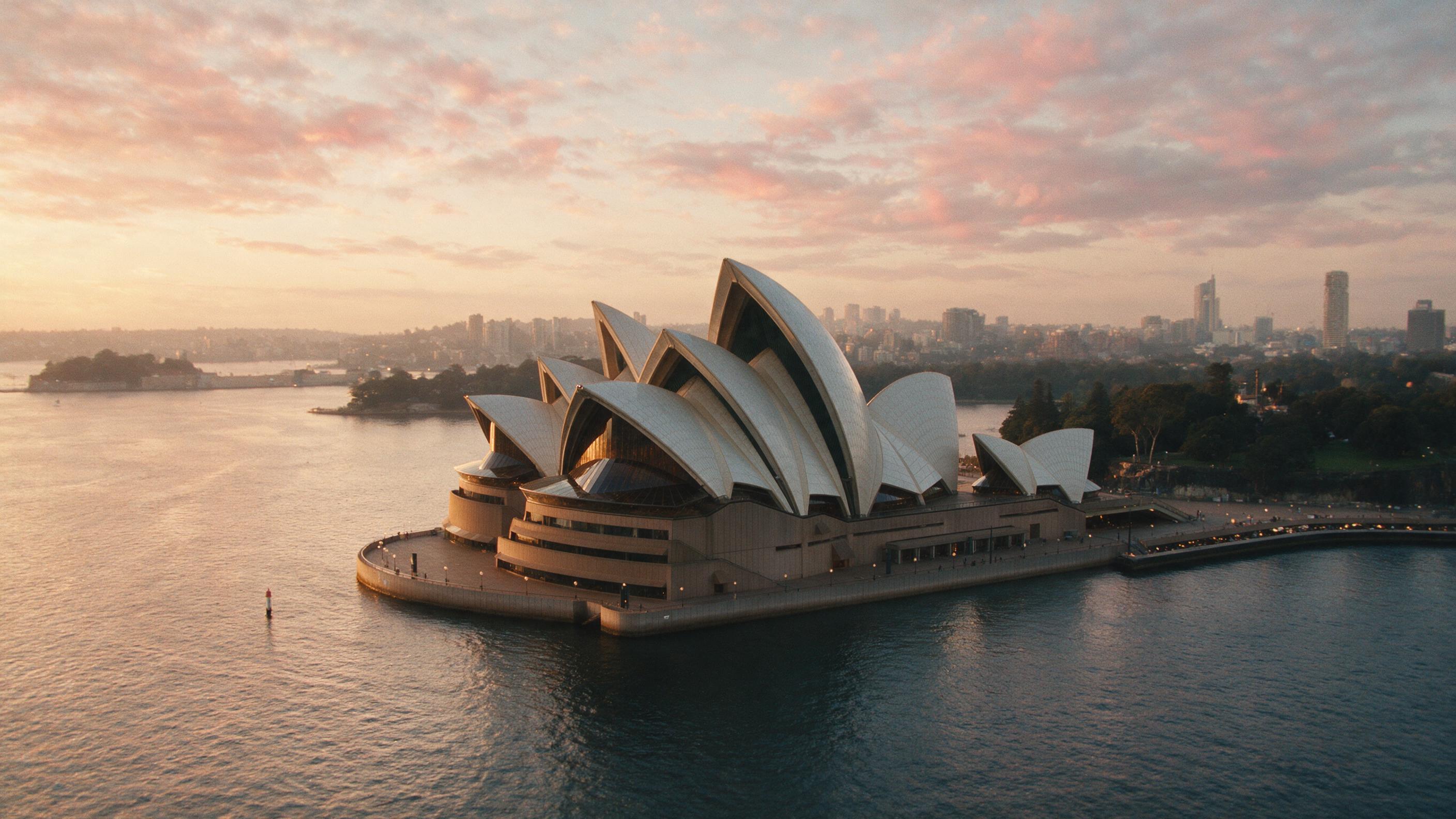 A sweeping aerial view of the Sydney Opera House at dusk, soft golden hour light reflecting off the harbor, clouds streaked pink and orange, distant city skyline blurred by slight haze, 35mm lens, natural grain, peaceful mood