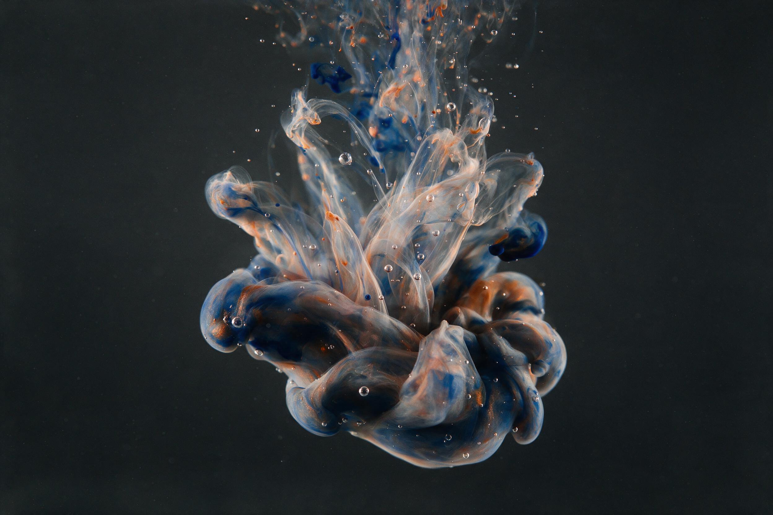 Fluid acrylic ink swirling in clear water, bursts of ultramarine and copper orange, delicate bubbles suspended mid-motion, backlit by a single soft studio light, dark charcoal background, minimal composition with negative space, subtle film grain.