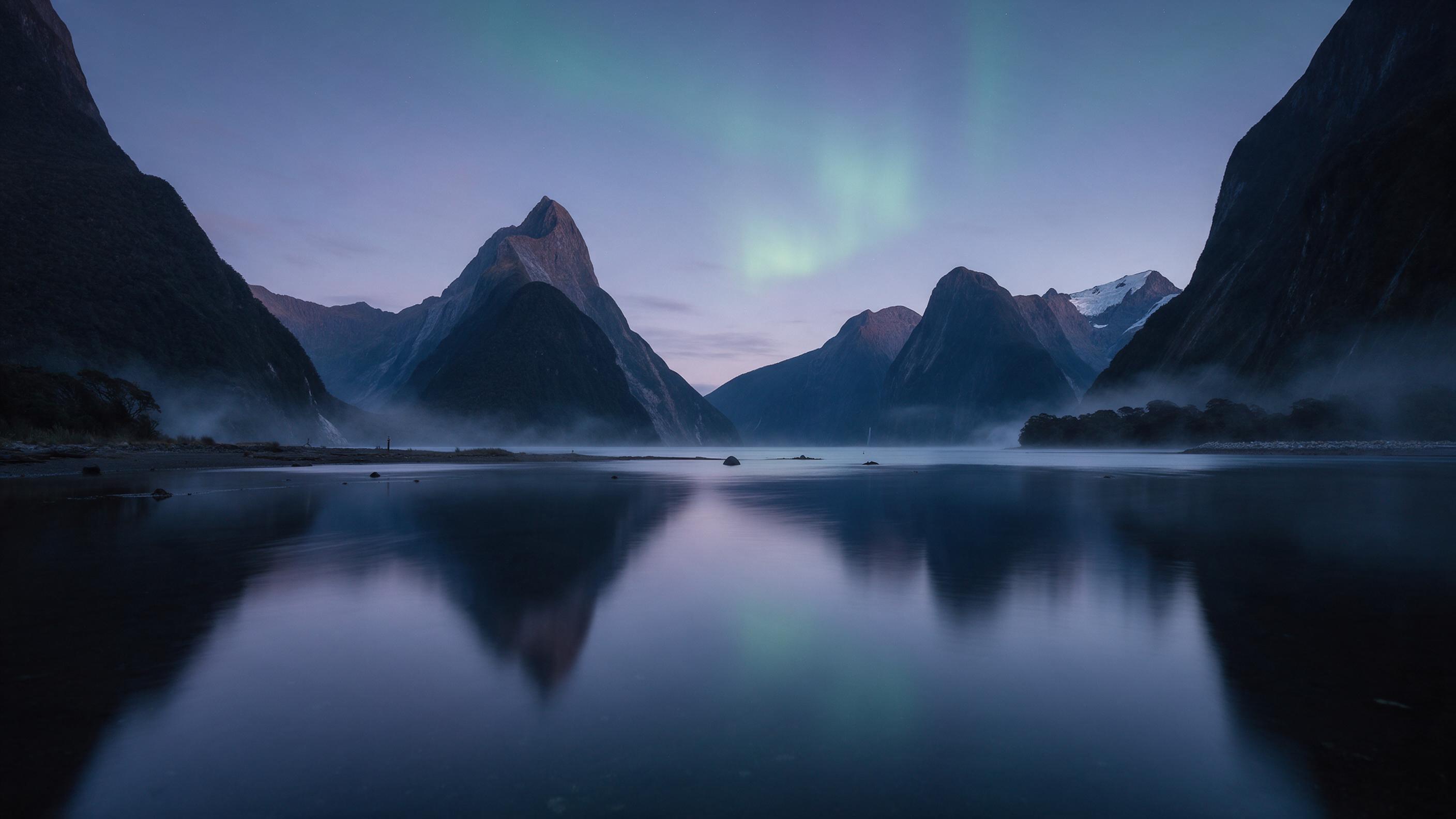 A glacial fjord at twilight, calm water reflecting sharp peaks with a faint green aurora overhead, thin mist curling at the shoreline, cold blue and purple tones, long exposure smoothness, minimal human presence