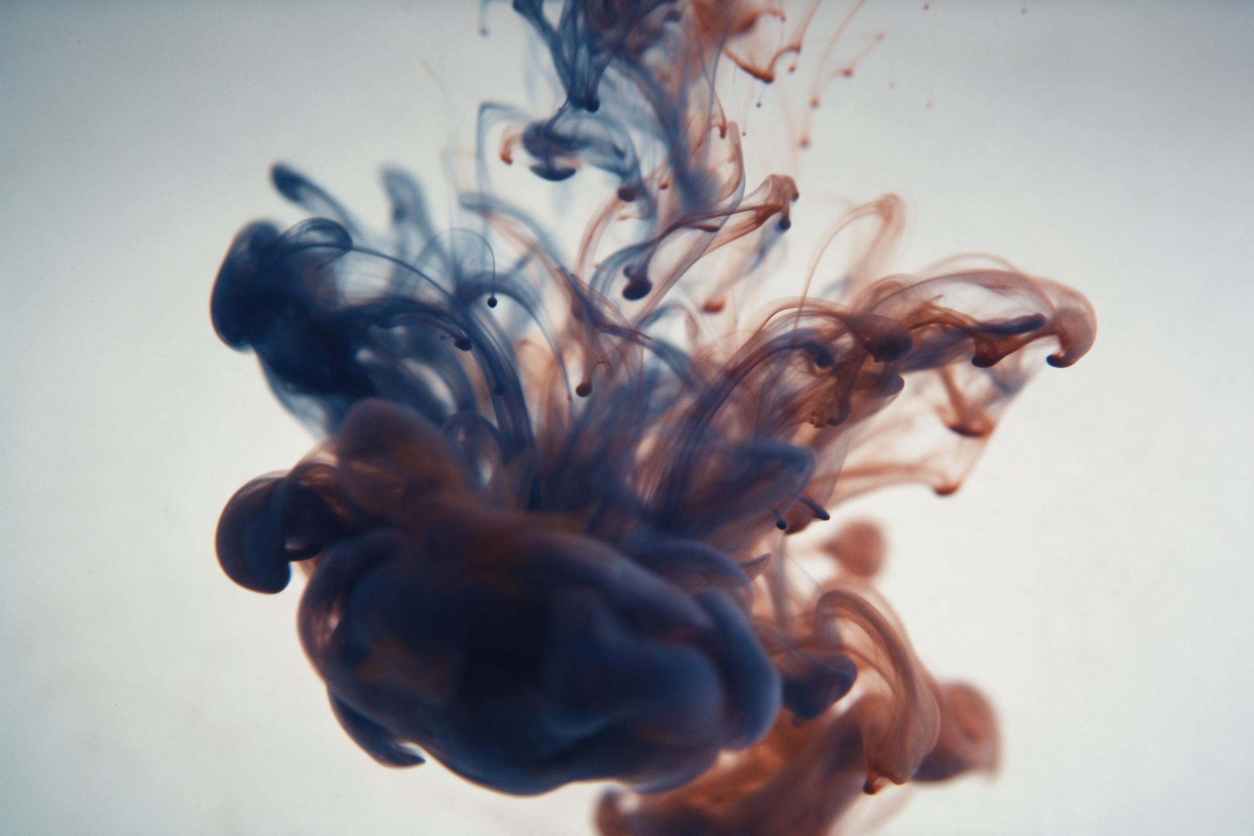 A macro view of swirling liquid ink in deep indigo and burnt sienna, dissolving into wispy tendrils of smoke, soft diffused backlighting, subtle film grain, minimal composition with negative space, no text, gentle motion blur conveying fluidity and quiet tension.