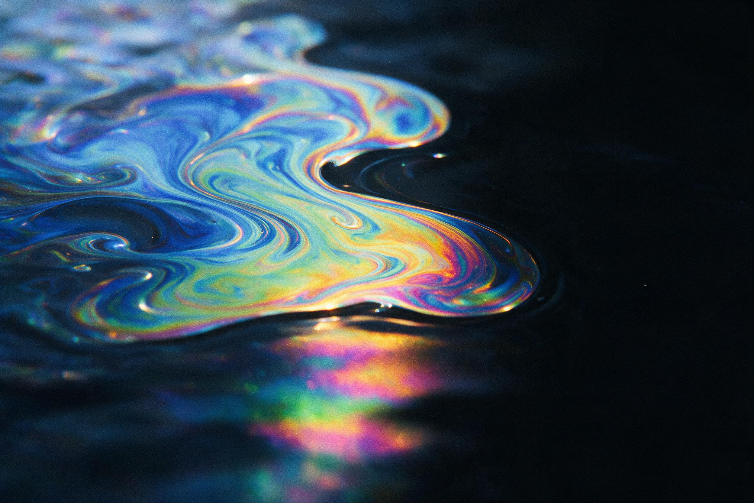 A sweeping macro shot of swirling iridescent oil film on water, fractured light creating fluid rainbow patterns, deep black background, soft reflections, subtle grain, balanced composition with negative space, minimalist surreal aesthetic