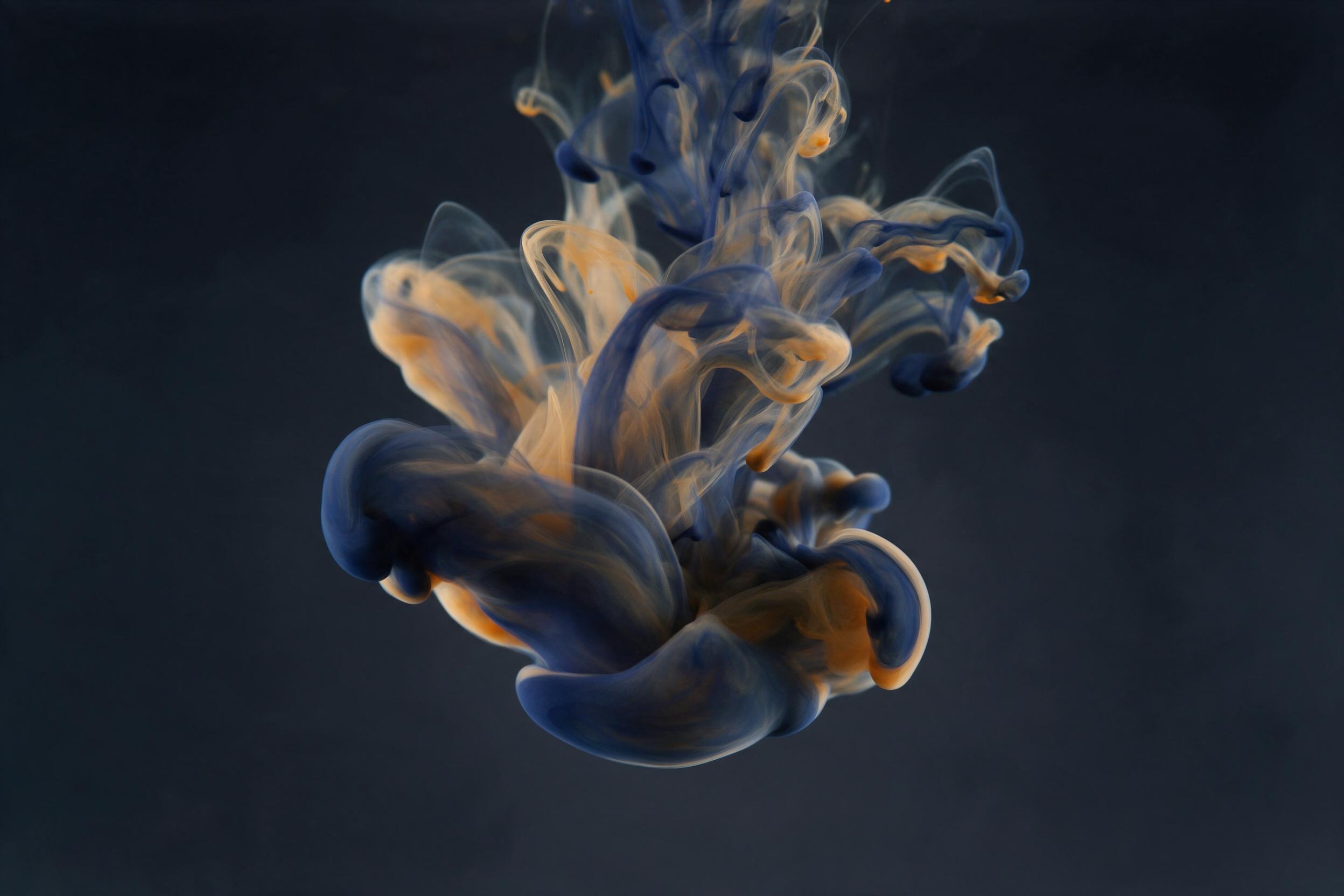 Fluid ink dissolving in clear water, swirling smoke-like tendrils, deep indigo merging with warm amber, dark background, soft studio lighting, macro shot, gentle motion blur, minimalist composition