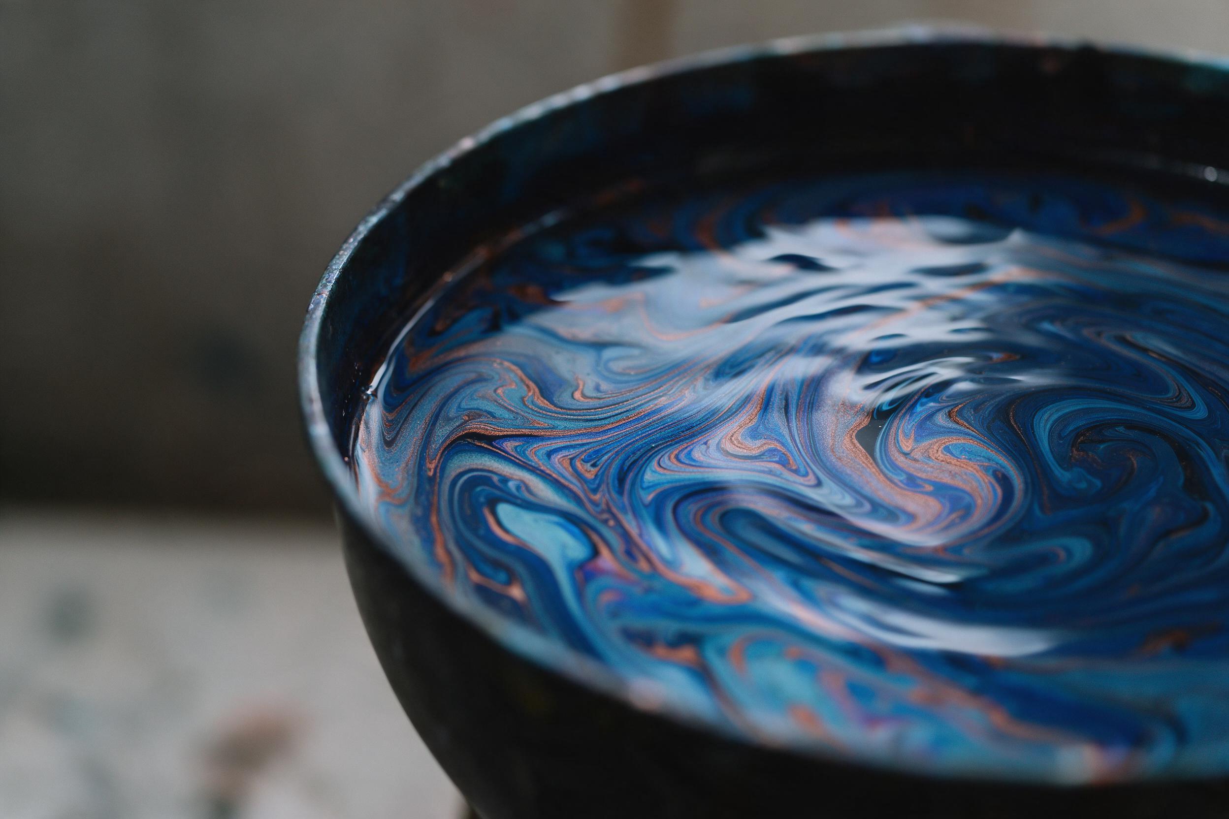 A close-up of fluid acrylic paint swirling in a dark basin, iridescent blues and copper veins intertwine, soft studio lighting from above creates gentle reflections, subtle ripples distort the pigment pattern, minimal composition with negative space on the left, smooth glossy texture, muted backgrou
