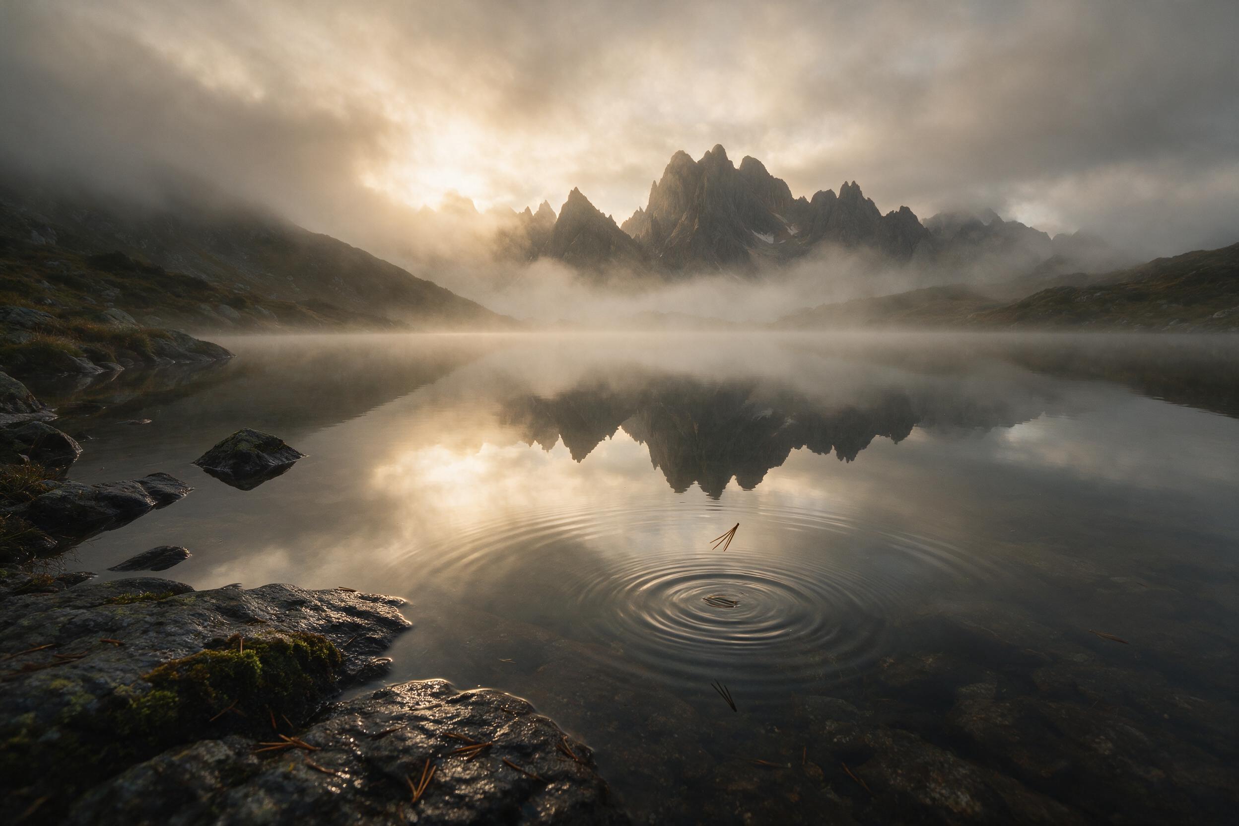 A misty alpine lake at dawn, mirror-like surface reflecting jagged peaks, soft golden light breaking through low clouds, foreground with wet rocks and moss, gentle ripples from a falling pine needle, muted earth tones, wide-angle composition emphasizing stillness, natural atmospheric depth.