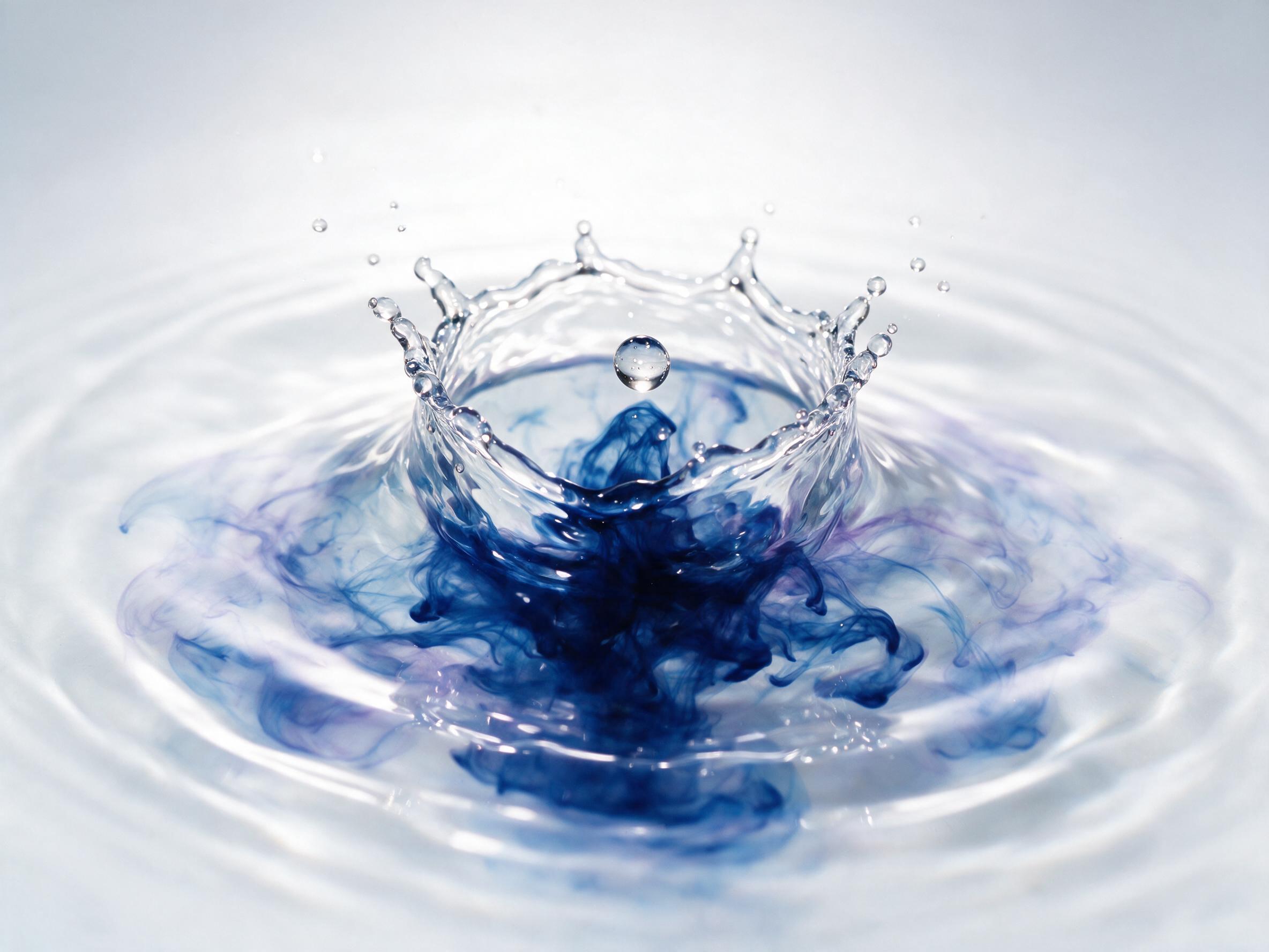 A single luminous droplet of deep blue ink falls into a pool of clear water, mid-splash, with delicate ripples and tendrils unfurling like smoke. Soft, diffused studio lighting from above creates a subtle gradient on the pure white background, emphasizing the droplet's sharp, mirrored surface and th