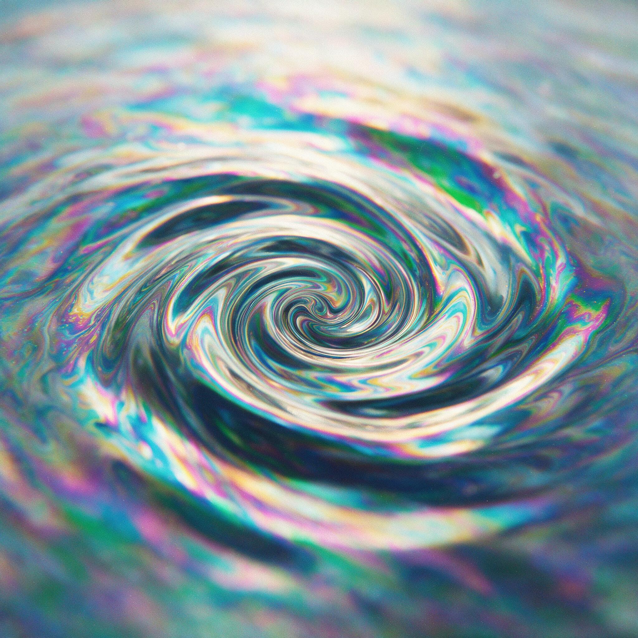 A swirling vortex of iridescent oil on water, vivid cyan and magenta reflections, soft diffused sunlight from above, shallow depth of field with blurred edges, slight grain, minimal composition focusing on the fluid movement and color blending.