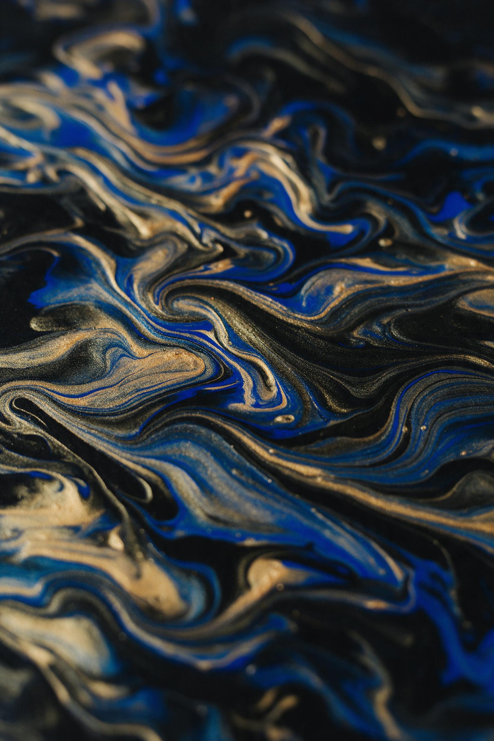 Fluid acrylic swirls of cobalt and gold on black canvas, macro shot capturing metallic pigment ridges, soft studio light raking across the textured surface, warm highlights on raised edges, cool shadows in deep valleys, shallow depth of field blurring the periphery