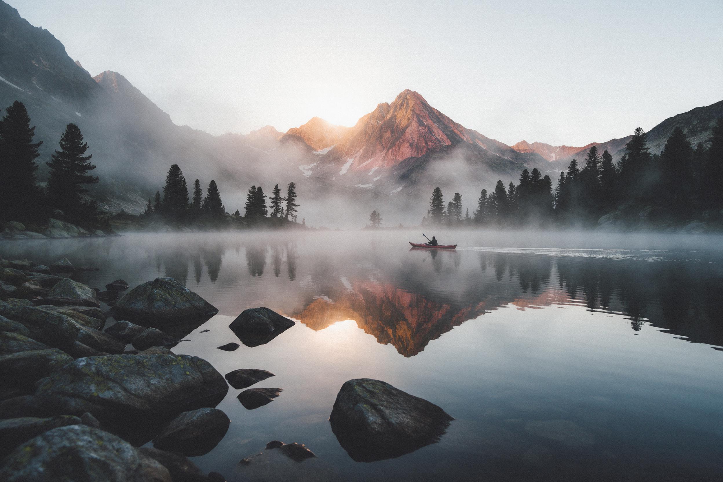 A misty alpine lake at dawn, mirrored peaks streaked with amber and rose, a lone kayak with a red hull cutting calm water, pine silhouettes lining the shore, soft fog drifting, warm light breaking over the ridge, slight grain, natural composition with leading lines from foreground rocks to the dista