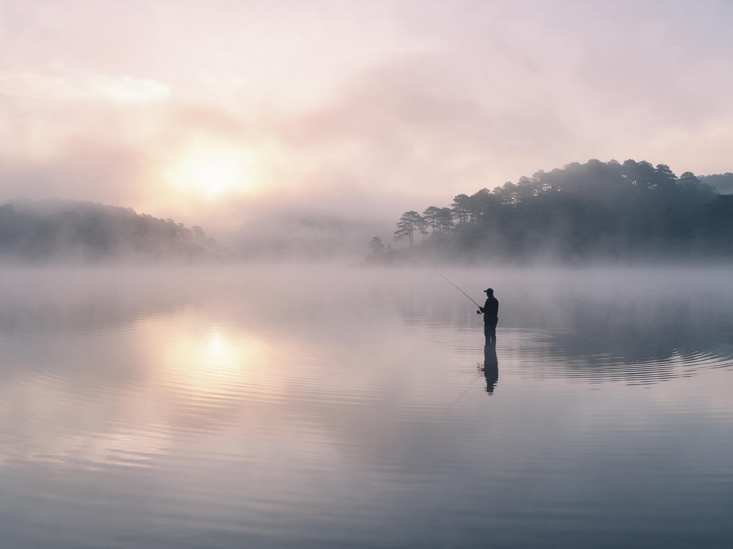 A lone fisherman stands on a mist-covered lake at dawn, silhouette against soft pink and gold sky, mirror-like water with gentle ripples, distant pine forest hills shrouded in fog, minimalist composition, airy atmosphere, muted color palette