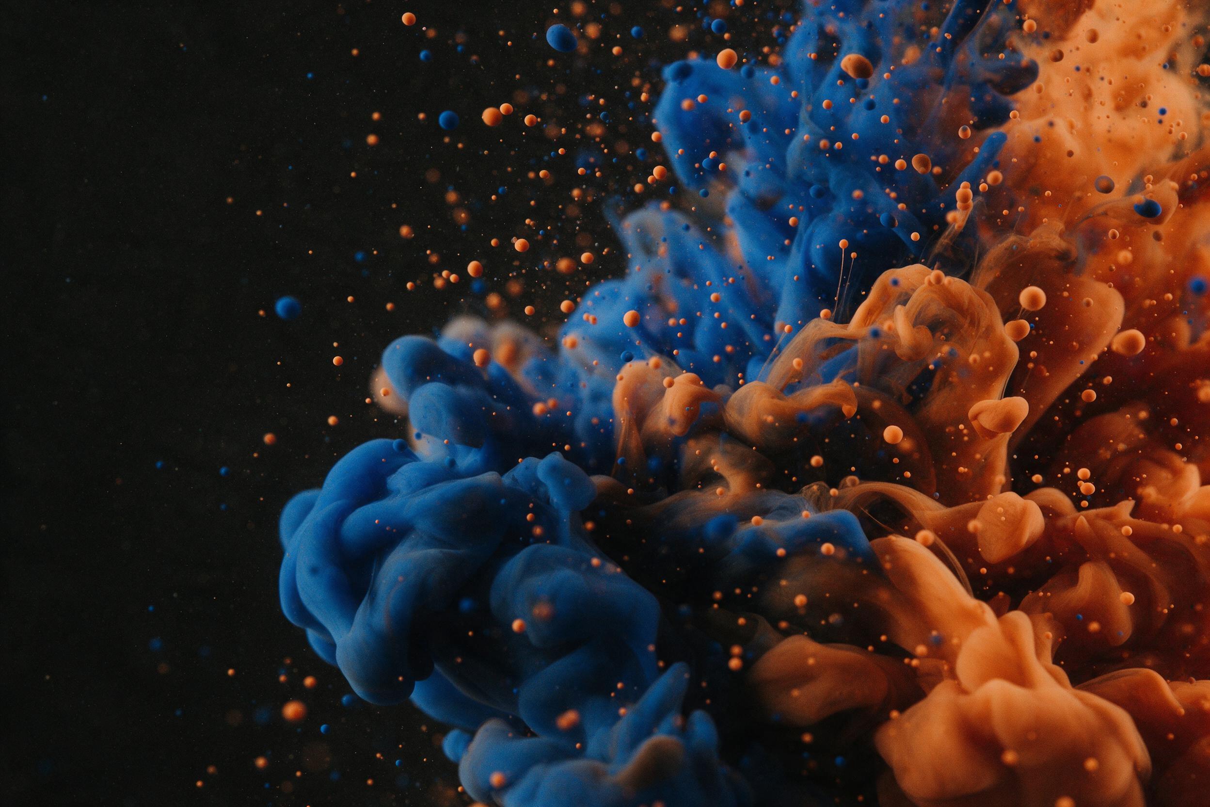 Fluid ink swirls of cobalt blue and burnt orange collide over a matte black canvas, captured with a macro lens. Fine droplets suspended mid-air catch warm side lighting, creating soft shadows. Composition balanced diagonally, negative space on the left. Grainy texture, slight motion blur.