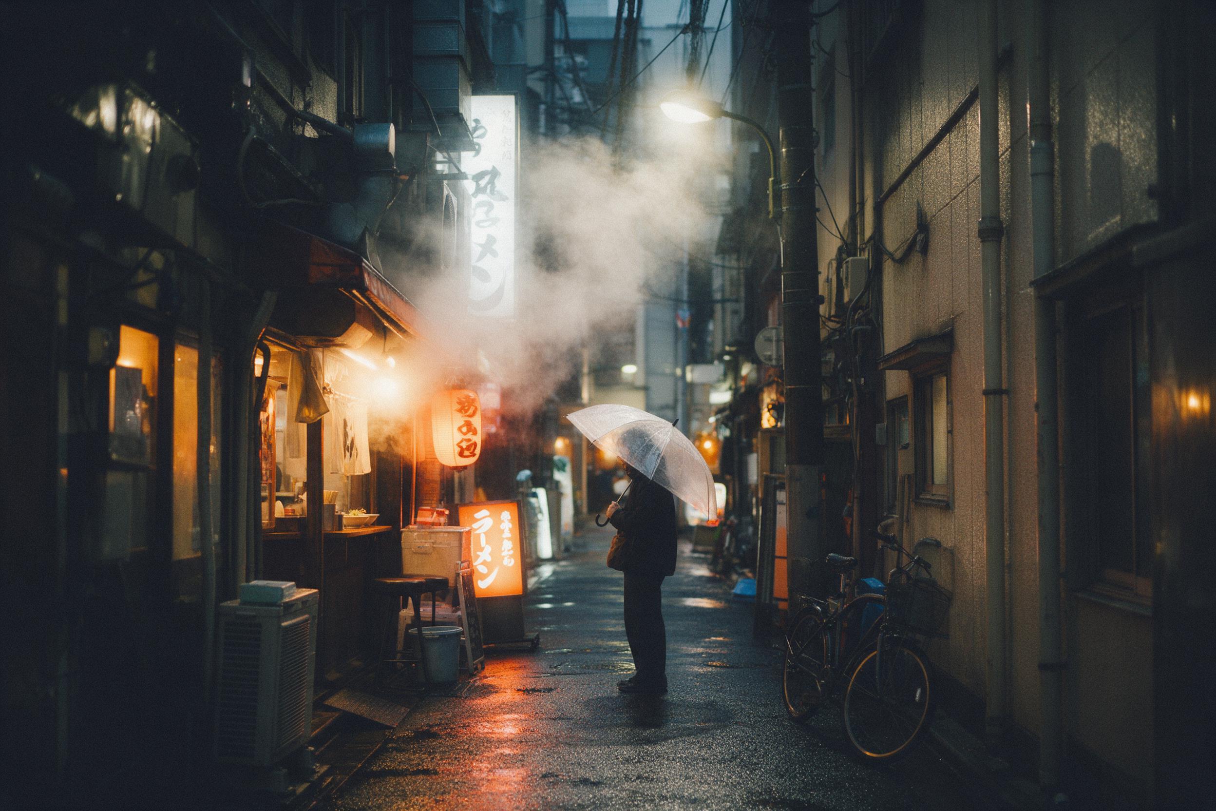 A cozy rainy evening in a narrow Tokyo alley, steam rising from a ramen shop, warm neon glow reflecting on wet asphalt, a lone pedestrian with a transparent umbrella under a soft streetlamp, shallow depth of field, natural grain, muted tones with orange accents
